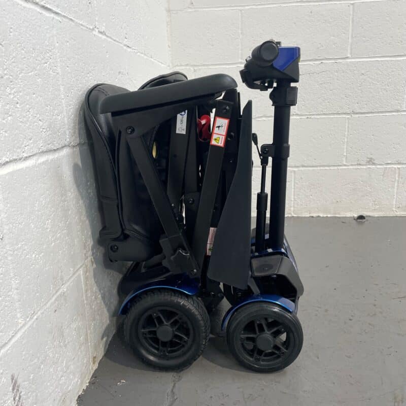 Side View of a Drive Flex Automatic Folding Mobility Scooter in Its Folded State, Placed Against a White Wall on a Gray Floor. the Scooter is Predominantly Black with Blue Accents on the Lower Body. It's Folded Down into a Compact Form, with the Seat Fully Collapsed Onto the Base and the Tiller Folded Down Alongside. the Black Wheels and Sturdy Frame Are Visible, with Warning Stickers and a Red Safety Lever Clearly Shown on the Side. This Image Highlights the Scooter's Convenience for Storage and Transport. Drive Flex Autofold with Carry Case & off Board Charger