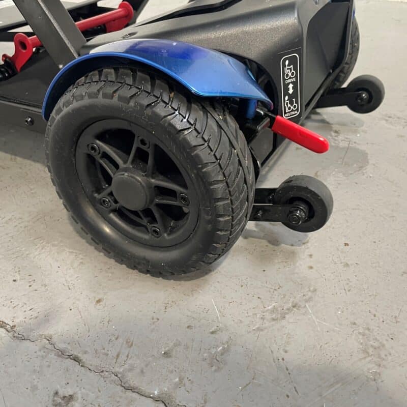 Close-up of the Rear Side of a Drive Flex Automatic Folding Mobility Scooter Showcasing Its Stability Wheels and Handbrake. the Scooter Features a Large Black Rear Wheel with a Robust Tread for Traction and a Smaller Anti-tip Stability Wheel Behind It. a Red Handbrake Lever is Mounted Near the Wheel for Easy Access and Safe Parking. Above the Handbrake, Instructional Icons Indicate the Driving Mode and Freewheel Options. the Scooter's Body is Black with a Blue Fender over the Main Wheel, Adding a Pop of Color to the Design. Drive Flex Autofold with Carry Case & off Board Charger