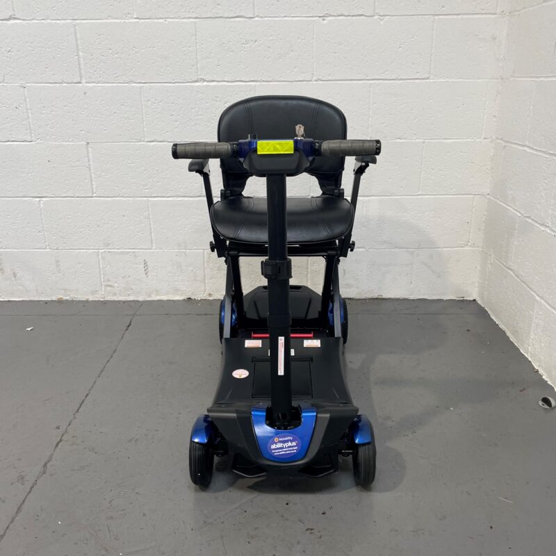 Front View of a Drive Flex Automatic Folding Mobility Scooter. the Scooter is Black with Blue Accents on the Base. the Tiller with the Control Panel is Folded Down Flat Against the Scooter's Vertical Support Column, with the Handlebars Resting Horizontally on Top, Featuring a Yellow Strip for Visibility. the Black Leatherette Captain's Seat with Armrests is Fully Upright. the Scooter's Compact Design is Highlighted by the Front Wheel Neatly Placed Between the Two Larger Back Wheels, Showcasing Its Portability and Space-saving Features. Drive Flex Autofold