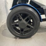 Close-up View of the Rear Wheel of a Drive Flex Automatic Folding Mobility Scooter. the Wheel is Made of Durable Black Plastic with a Five-spoke Design, and It's Fitted with a Solid Rubber Tire for Traction and Stability. a Blue Trim Accentuates the Edge of the Wheel, Matching the Scooter's Color Scheme. the Wheel is Attached to the Scooter's Frame, Which Has a Matte Black Finish. Drive Flex Autofold
