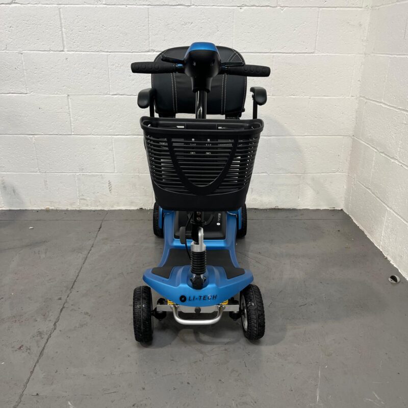 Front View of a Li-tech Neon 15 Mobility Scooter. the Scooter Has a Blue and Black Color Scheme, with a Prominent Black Wire Basket on the Front. Attached to the Basket is the Blue Tiller with the Scooter's Controls. the Scooter Features a High-backed, Black Seat with Armrests, and a Black Steering Column with Ergonomic Handlebar Grips. the Front Wheel is Connected to Yellow Suspension Springs Visible Under the Blue Body of the Scooter, Which Sits on Solid Black Tires with Silver Rims. Li-tech Neon 15