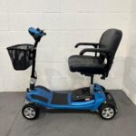 Left-hand Side View of a Li-tech Neon 15 Mobility Scooter Parked Indoors. the Scooter Has a Sleek Black and Blue Design with a High-backed, Black Leatherette Seat with Armrests. the Seat is Mounted Above a Blue Chassis with Black Anti-slip Foot Mats. Silver Rims on the Tires and a Front Wheel with Yellow Suspension Springs Are Visible, Indicating a Focus on Comfort and Stability. a Black Wire Basket is Attached to the Blue Tiller, Which Also Has a Simple Control Panel. Li-tech Neon 15